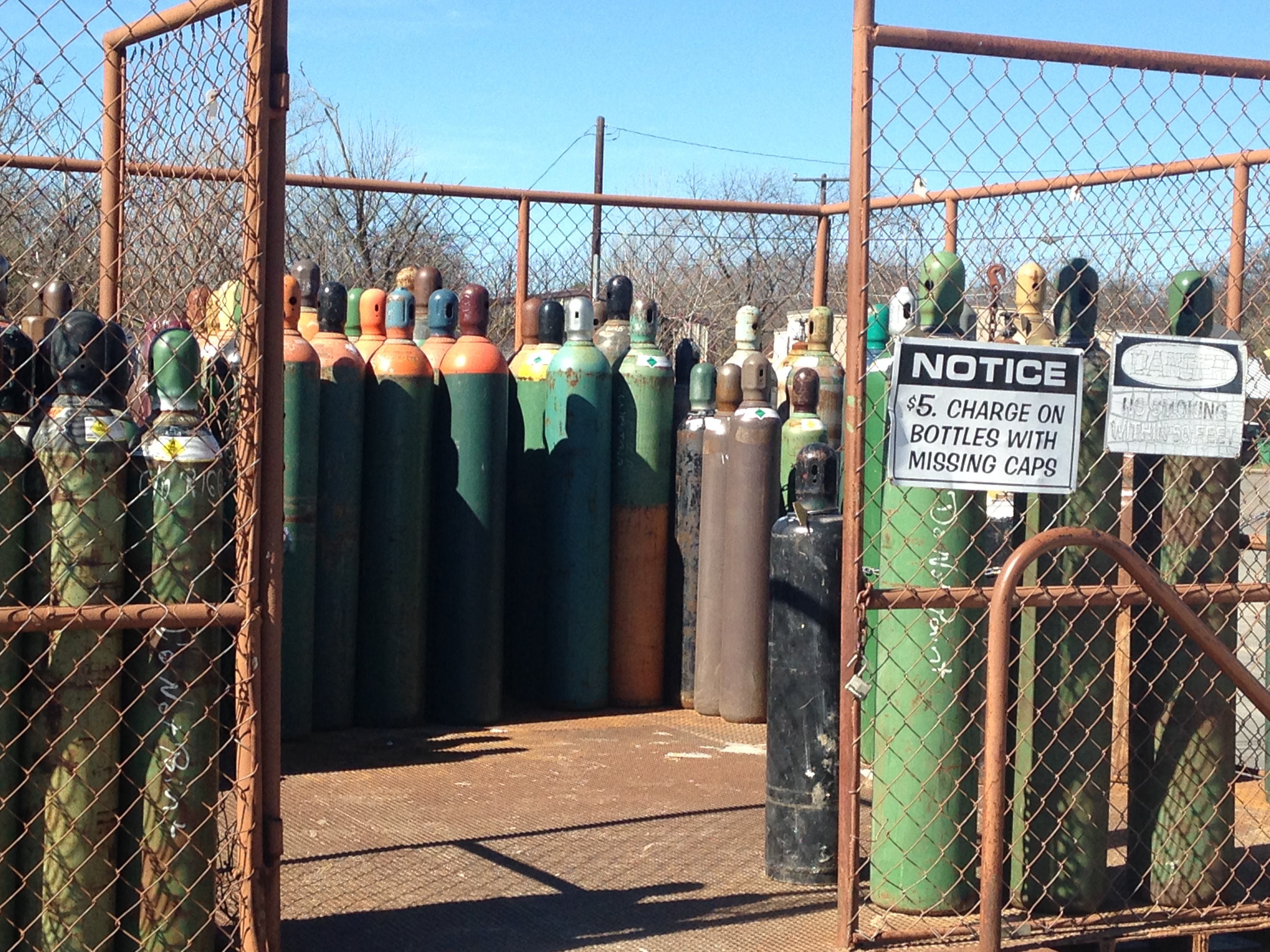 Outdoor gas cylinder exchange yard at Riggs Machine & Welding in Stephenville, TX — rows of green, orange, and black welding gas bottles behind a chain-link fence