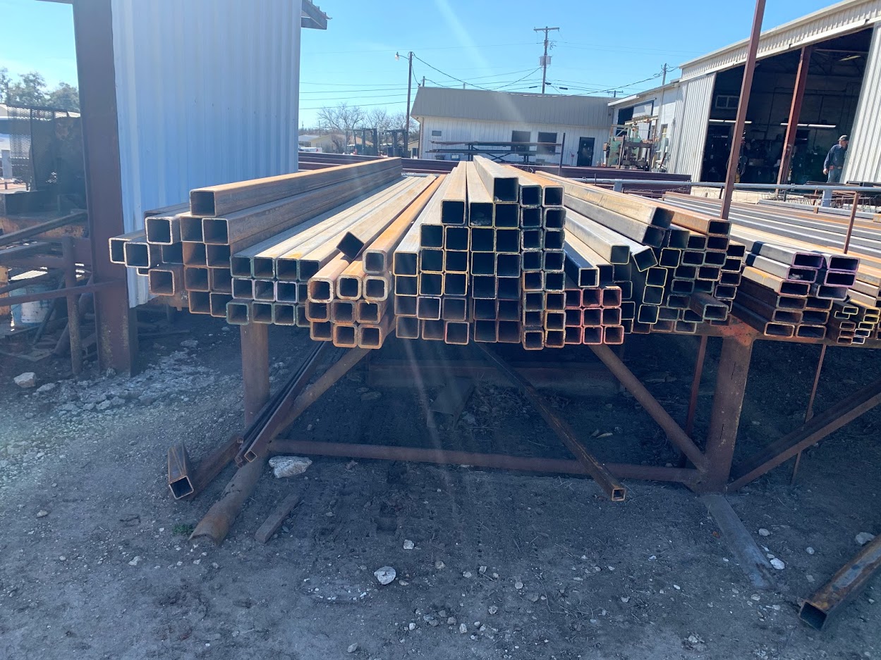 Heavy quantity of square steel tubing stacked at Riggs Machine & Welding in Stephenville, TX with shop building behind