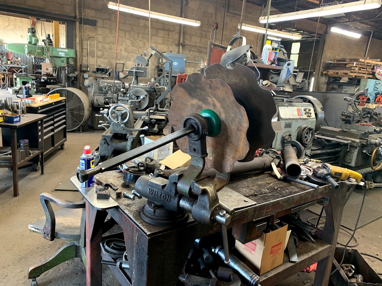 Riggs Machine & Welding shop floor in Stephenville, TX — Wilton vise with a large gear shaft in the foreground and rows of lathes and milling machines behind