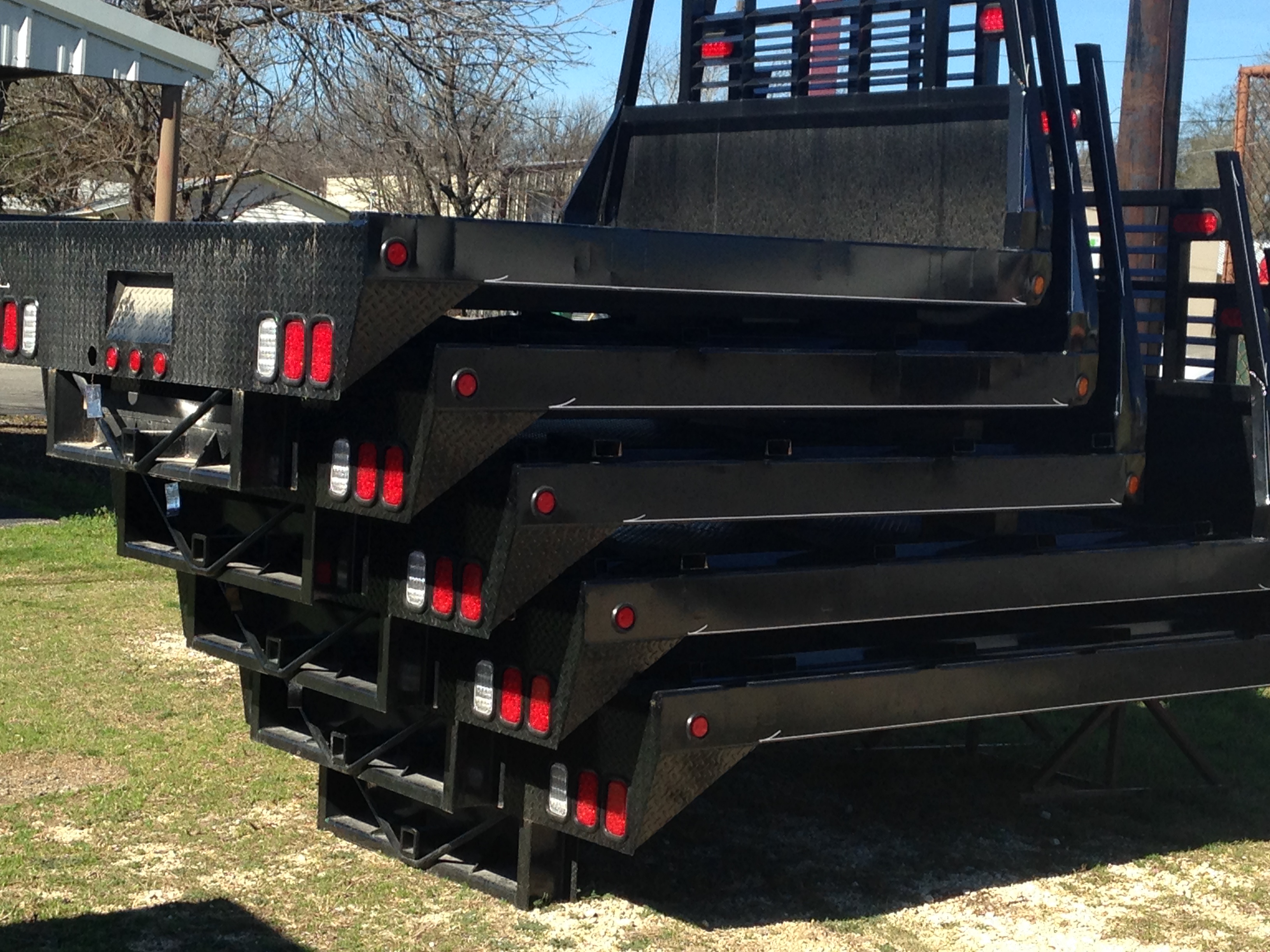 Multiple black J-I MFG flatbed truck beds stacked at Riggs Machine & Welding in Stephenville, TX, showing tailgates, LED lights, and checker plate decking